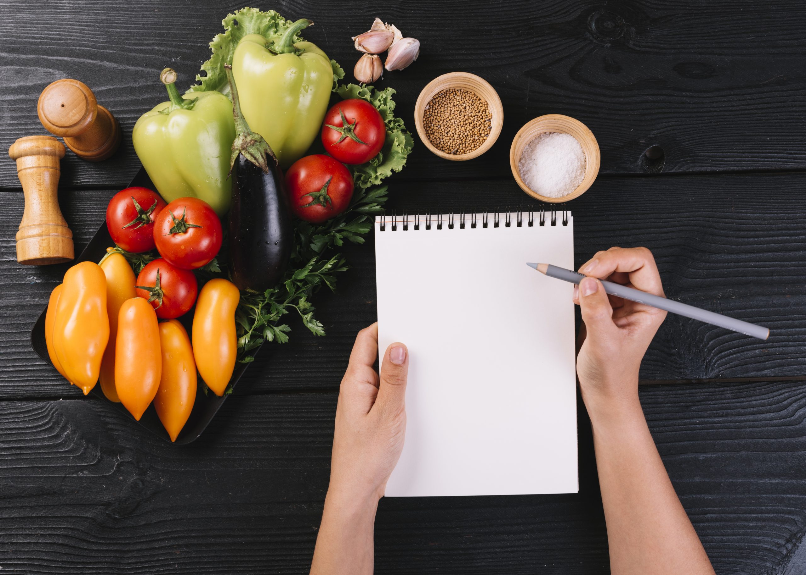 person s hand writing spiral notepad near vegetables spices black wooden surface scaled